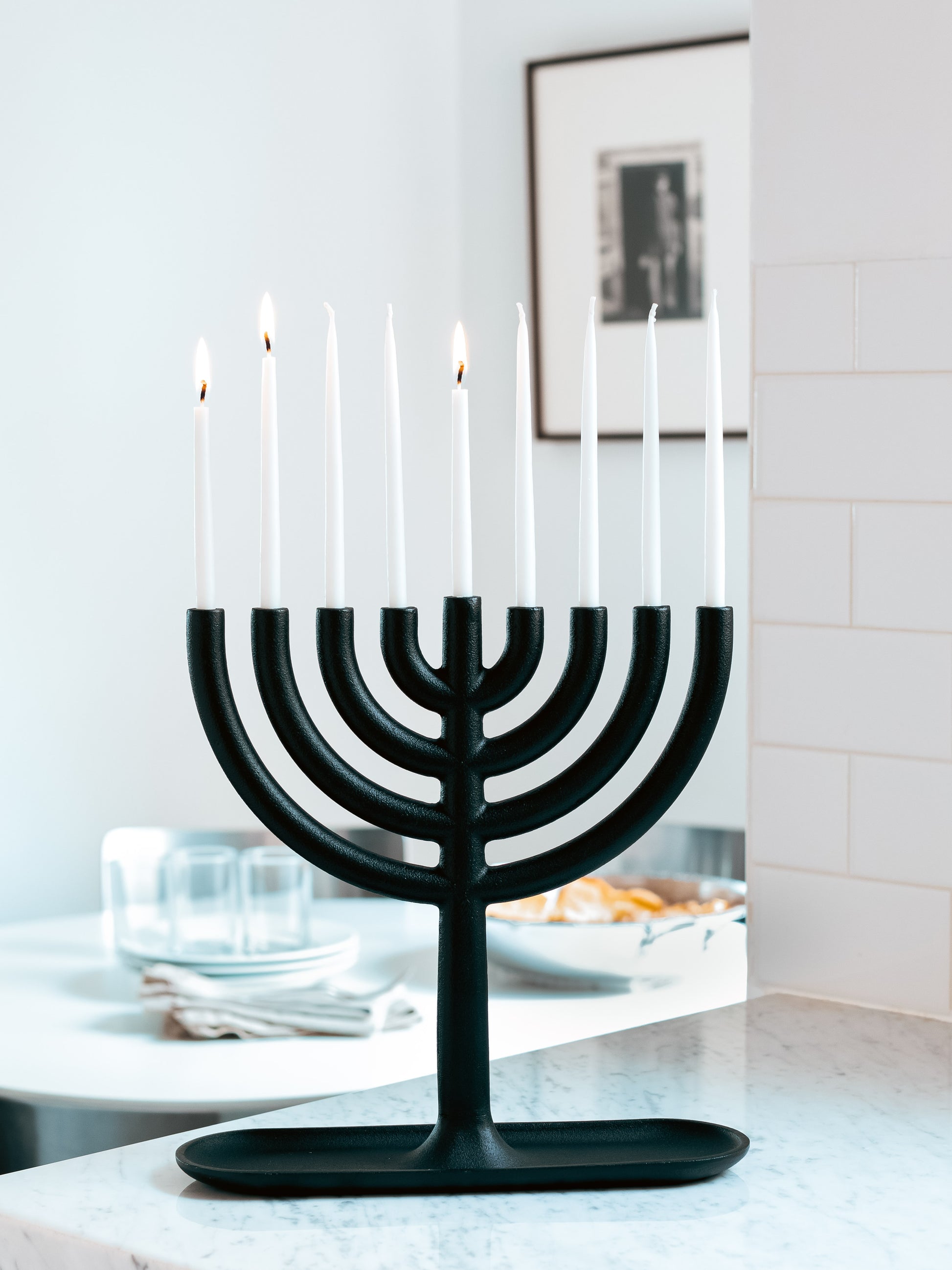Black cast-iron Menorah with four lit and five unlit white taper candles on a marble counter.