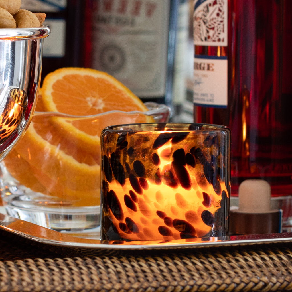 Tortoise votive glass candle holder with a lit candle, next to orange slices and a silver bowl.