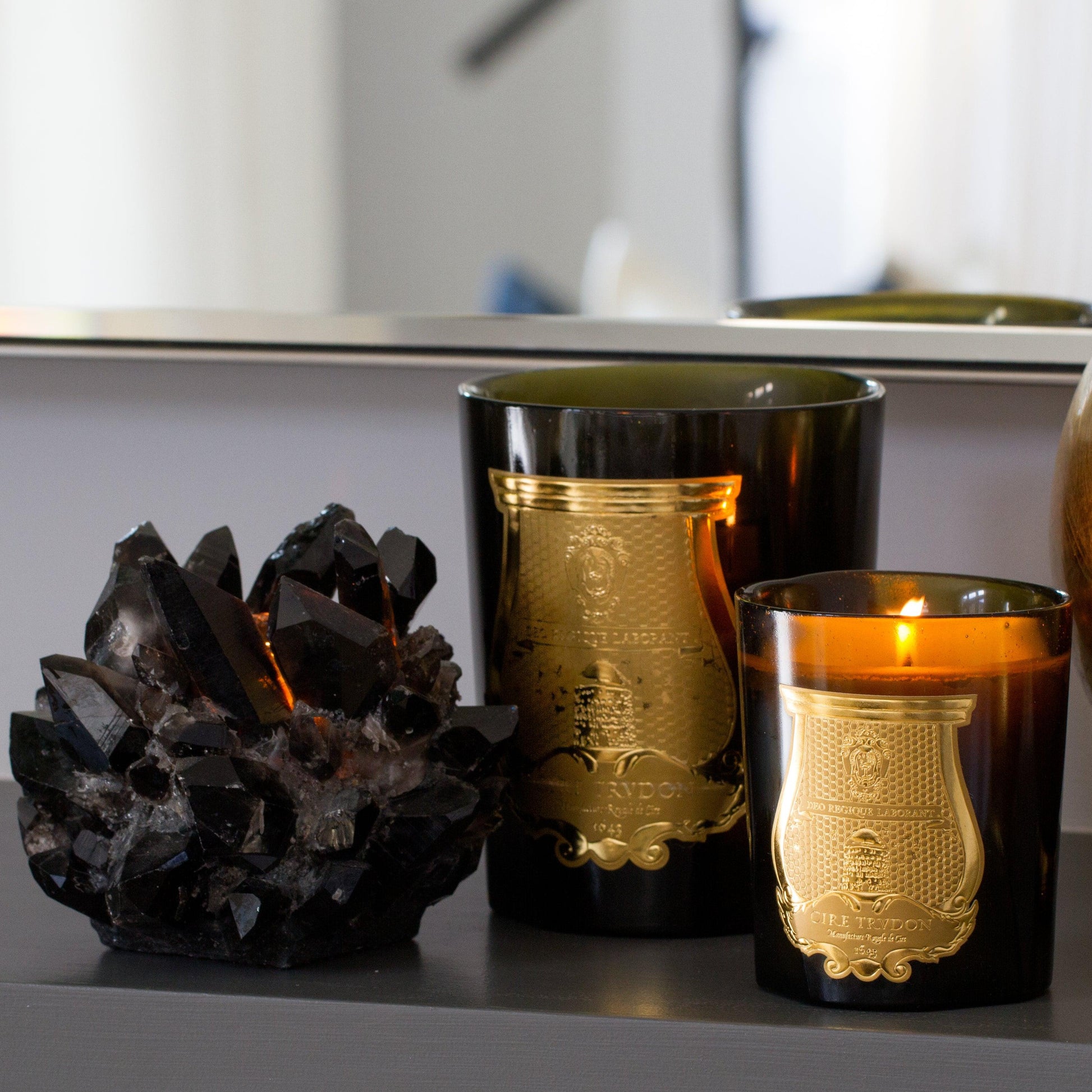 Two Trudon Abd El Kader scented candles in dark green glass vessels with gold labels, one lit, next to a dark crystal cluster.