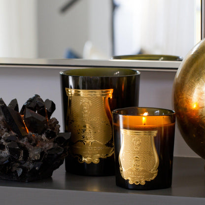 Trudon Spiritus Sancti scented candles in dark glass vessels with gold labels, one lit, next to a black crystal.
