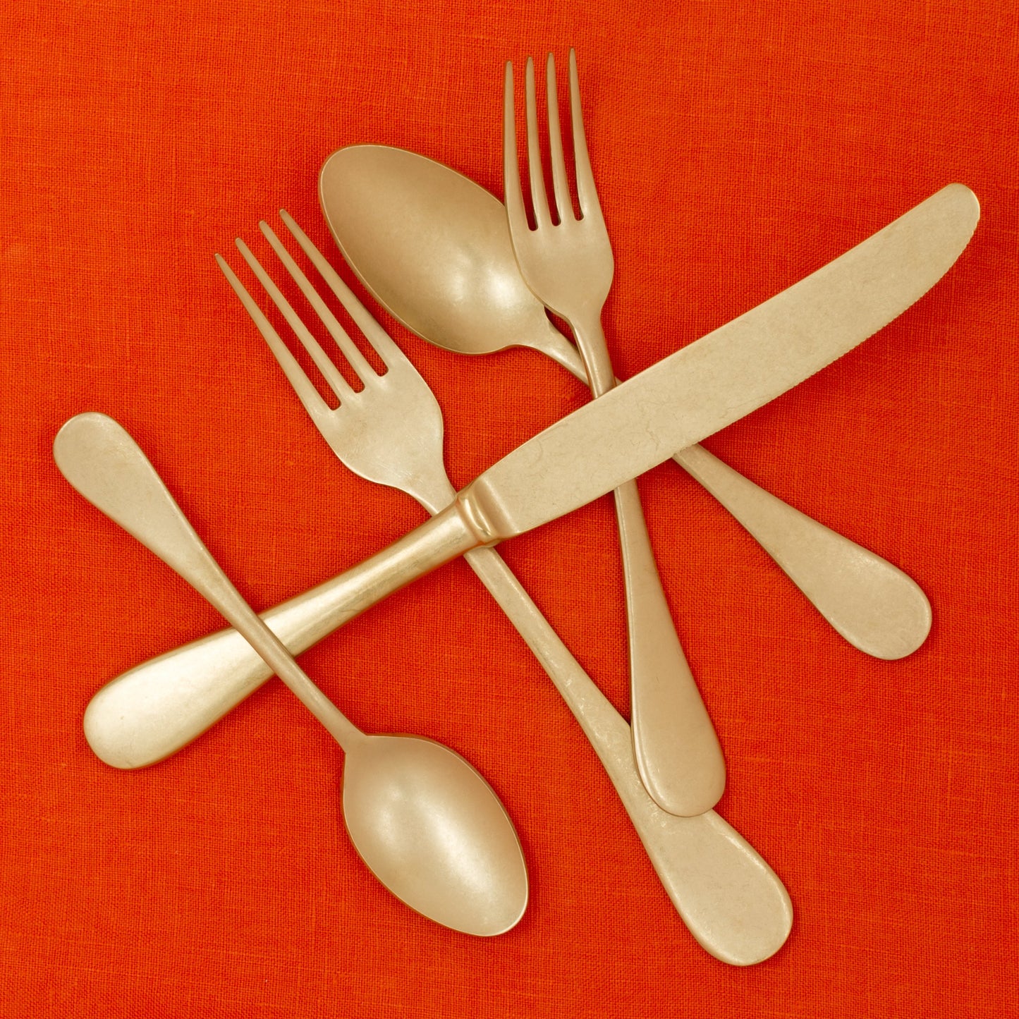 Vintage Oro 5-piece gold-finish stainless steel flatware set on an orange linen background.