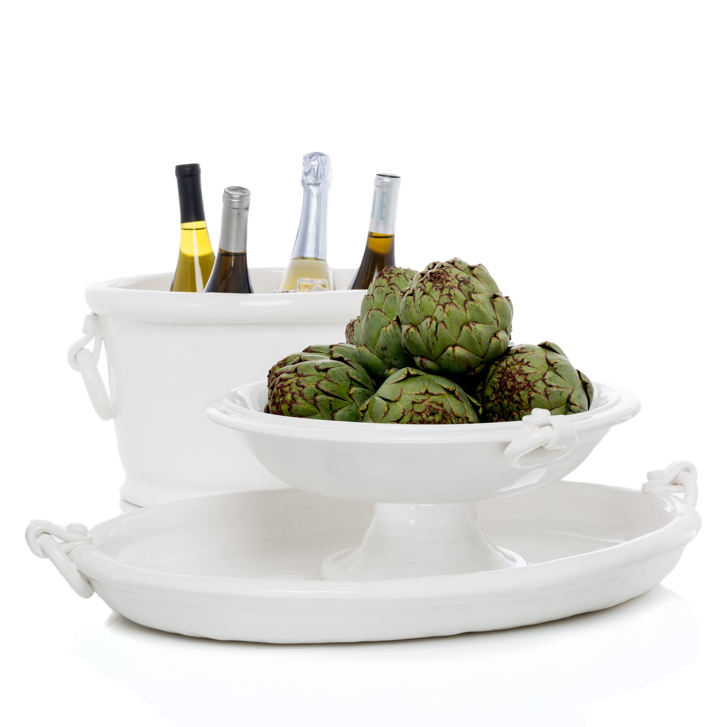 White ceramic oval ice bucket with ring handles, holding wine bottles, beside a matching serving bowl and tray with artichokes.