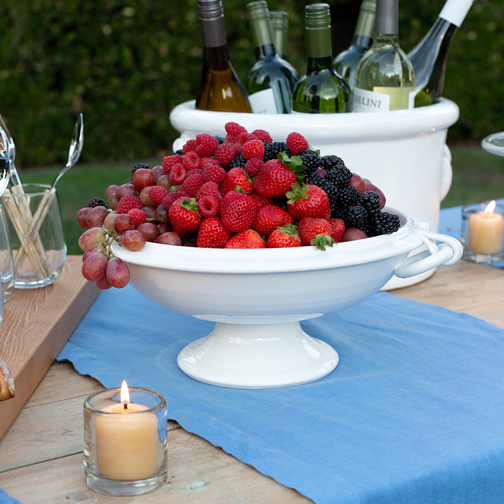 White ceramic pedestal fruit bowl with ring handles, filled with berries and grapes, on a wooden table.