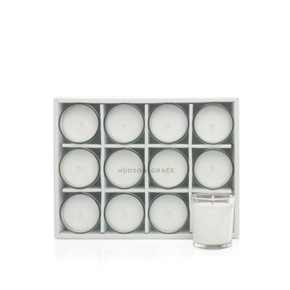 White paraffin votive candles in clear glass cups, set of 12, with one lit candle.