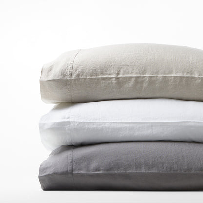 Stack of three Washed-Linen Pillowcases: light beige, white, and charcoal gray, with double-needle stitching.